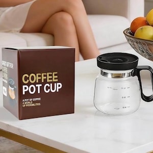 May include: A clear glass coffee pot cup with a black lid and handle sits on a white table next to a brown box. The box reads "COFFEE POT CUP" in gold lettering. A bowl of fruit is in the background.