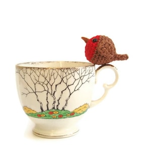 May include: A cream-colored teacup with a gold rim and a decorative tree and landscape design. A crocheted bird with a red breast and brown body sits on the cup's handle. The teacup has a pedestal base.