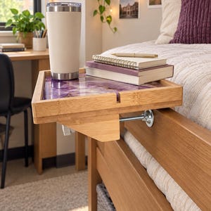 May include: A bamboo bedside tray with a purple marbled resin inlay, featuring a metal clamp mechanism for attachment to a bed frame. The tray holds a tall beige insulated tumbler, a spiral-bound notebook, and a pen, providing a flat surface.