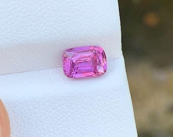 Può includere: Una gemma rosa vibrante, tagliata a cuscino con una superficie sfaccettata, che mostra tonalità magenta profondo e rosa più chiaro. La pietra è posizionata su un cartoncino bianco testurizzato, che ne evidenzia la chiarezza e la precisione.