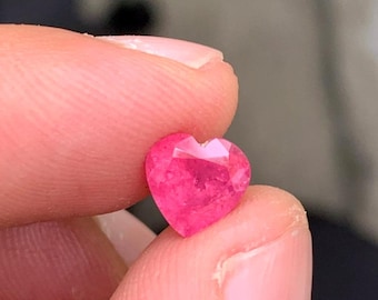 Può includere: Una gemma rosa acceso tagliata a forma di cuore con molteplici sfaccettature che riflettono la luce. La pietra presenta una base appuntita e una parte superiore incavata, mostrando un colore profondo e saturo in tutta la sua struttura.