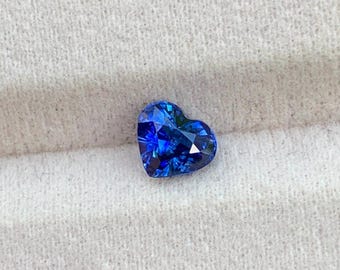 Può includere: Una pietra preziosa a forma di cuore di un colore blu profondo e vibrante con una superficie sfaccettata che riflette la luce. La pietra presenta una base appuntita e una parte superiore arrotondata, con un taglio brillante che esalta la tonalità.