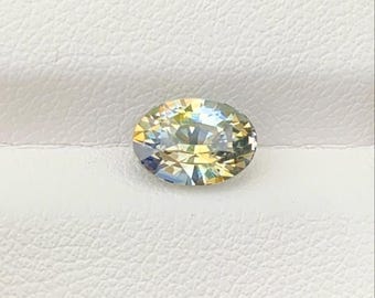 Può includere: Una pietra preziosa dal taglio ovale con una superficie sfaccettata che mostra uno spettro di colori iridescenti, inclusi riflessi di luce gialla, blu e bianca. La pietra è posizionata su una superficie bianca testurizzata con grana.