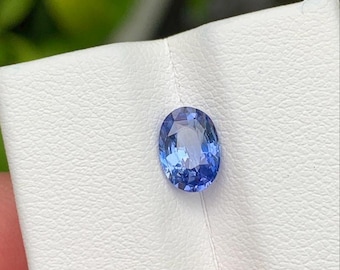 Può includere: Una pietra preziosa blu dal taglio ovale con superficie sfaccettata, che mostra varie tonalità di blu chiaro e scuro. La pietra è posizionata su un cartoncino bianco testurizzato, che ne evidenzia la trasparenza e l'aspetto lucido.