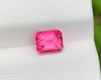 Puede incluir: Una gema de color rosa intenso tallada en forma de esmeralda rectangular con superficies facetadas que reflejan la luz. La gema está colocada sobre una superficie de exhibición blanca y texturizada, mostrando su color intenso y estructura.