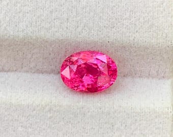 Puede incluir: Una gema de corte ovalado con un color rosa intenso, profundo y saturado, que presenta múltiples facetas que reflejan la luz. La piedra se muestra descansando sobre un inserto de terciopelo blanco texturizado.