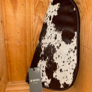 May include: A teardrop-shaped bag featuring a brown and white cowhide pattern with a dark brown leather trim. A rectangular grey tag is attached to the bag, displaying the text MYRA and a website address in white lettering near the bottom.