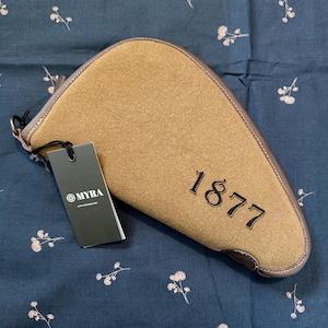 May include: A saddle-shaped tan canvas pouch with brown leather trim and the number 1877 embroidered in black. A black rectangular tag attached to the side features the white text MYRA and a circular logo. The pouch has a zipper closure.