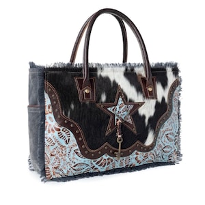 May include: A tote bag featuring black and white cowhide leather, dark brown leather trim, and light blue floral embossed leather panels. The bag includes a central star cutout, metal studs, a hanging key charm, and frayed edges on the sides.