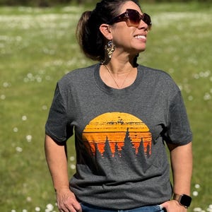 May include: Dark gray t-shirt featuring a sunset design with orange and yellow hues above a silhouette of trees. The shirt has a crew neck and short sleeves. The person is wearing sunglasses and blue jeans.