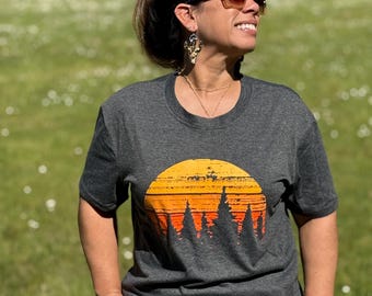 Puede incluir: Camiseta gris oscuro con un diseño de puesta de sol en tonos naranja y amarillo sobre una silueta de árboles. La camiseta tiene cuello redondo y mangas cortas. La persona lleva gafas de sol y vaqueros azules.