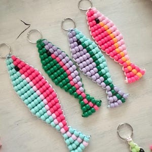 Puede incluir: Cuatro llaveros de peces de cuentas en tonos rosa, verde, morado y turquesa. Cada pez tiene un llavero plateado adjunto. Los peces están hechos de pequeñas cuentas de colores y están dispuestos sobre una superficie clara.