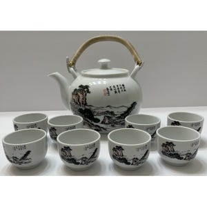 May include: A white ceramic tea set consisting of a teapot with a curved handle and seven matching cups. The teapot and cups feature black and light pink ink-style landscape illustrations of mountains and trees, along with black Chinese calligraphy.
