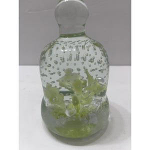 Puede incluir: Un pisapapeles de vidrio transparente con una base redondeada y una parte superior bulbosa. El interior presenta un diseño floral verde claro y numerosas burbujas de aire. El vidrio tiene un ligero tinte verde, y la forma general se asemeja a una botella.