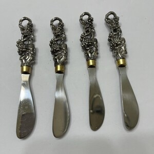 May include: A set of four silver-toned metal spreaders with textured handles featuring a grape cluster design. Each spreader has a smooth, flat blade and a small gold-toned metal band connecting the decorative handle to the stainless steel blade.