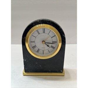 May include: A black marble quartz clock with a gold-tone bezel and base. The clock face is white with black Roman numerals and gold hands. The word "QUARTZ" is printed on the clock face.