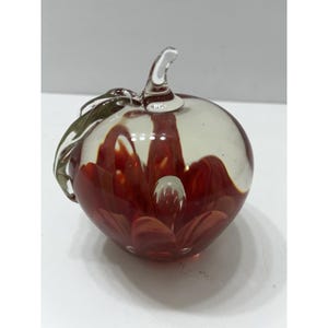 Puede incluir: Una escultura de manzana de cristal transparente con detalles en rojo y blanco. La manzana tiene un tallo y una hoja. El interior presenta un diseño floral rojo. La escultura está sobre una superficie blanca.