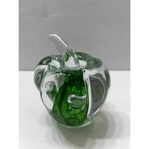 Puede incluir: Una escultura de manzana de vidrio transparente con un diseño de remolino verde en el interior. La manzana tiene un tallo y una forma redondeada. La escultura está sobre una superficie blanca.