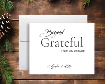 May include: A white greeting card with the words "Beyond Grateful" in elegant script, accompanied by "Thank you so much!" and a heart symbol with names. The card is placed with a white envelope on a wooden surface, decorated with pine and pinecones.
