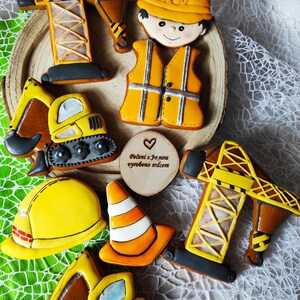 Könnte beinhalten: Eine Sammlung von Keksen zum Thema Bau, darunter ein Kran, ein Bagger, ein Schutzhelm, ein Verkehrskegel und ein Arbeiter. Die Kekse sind mit gelbem, orangefarbenem, braunem und schwarzem Zuckerguss verziert. Eine Holzscheibe mit Text ist ebenfalls vorhanden.