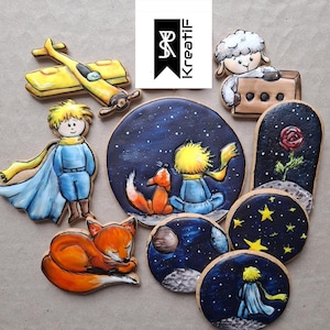May include: A selection of decorated biscuits depicting scenes and characters from "The Little Prince." Includes a yellow aeroplane, a boy in blue, a sleeping fox, and a sheep. The biscuits are decorated with blue, yellow, orange, and white icing.