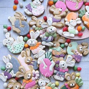 May include: A collection of Easter-themed sugar cookies shaped like bunnies, eggs, and carrots. The cookies are decorated with pastel colors, including pink, blue, green, and orange. Some bunnies are in carriages, and others are holding eggs. Colorful candies are scattered around the cookies.