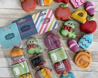 Peut inclure: Assortiment de biscuits décorés sur le thème de l'école, avec des formes de pommes, de crayons et de cahiers. Les biscuits sont emballés individuellement et disposés sur une surface imitation bois clair. Certains biscuits portent les mots "glue" ou "ABC".