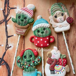 Puede incluir: Una colección de galletas navideñas festivas en palitos, con diseños de cactus y llamas. Las galletas están decoradas con glaseado colorido, incluyendo sombreros, luces y una bufanda. Una galleta tiene el texto "Merry Christmas".