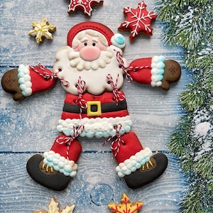 May include: A Christmas-themed biscuit arrangement featuring a Santa Claus figure and snowflake biscuits. The Santa biscuit has a red suit, white beard, and black boots. The biscuits are on a blue wooden surface, with a snowy pine branch in the background.