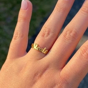 Può includere: Un anello dorato con il nome "Neale" scritto in corsivo. L'anello è indossato su un dito, con smalto rosa chiaro. Lo sfondo è sfocato, suggerendo un ambiente esterno.