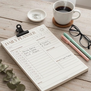 May include: A daily planner notepad with a black clip, featuring a schedule and goal section. A cup of coffee, pens, glasses, and a small dish with paper clips are on a wooden surface. The planner has the text "DAILY PLANNER".
