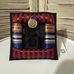 May include: A square, black fabric case with a burgundy, ribbed border. The case features colorful, patterned bands in blue, orange, and silver. A gold-colored medallion hangs from the top, and the case is set against a white surface.