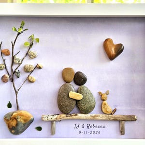 May include: A framed pebble art piece featuring two figures sitting on a driftwood bench with a small dog, a tree made of twigs and pebbles, and a heart-shaped stone. The text TJ & Rebecca 9-11-2026 is printed on a light purple background.