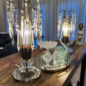 May include: Two vintage crystal lamps with cascading crystal accents and a brass-toned frame. The lamps sit on a wooden surface, with a mirrored tray holding decorative glass pieces. The lamps are lit, casting a warm glow.
