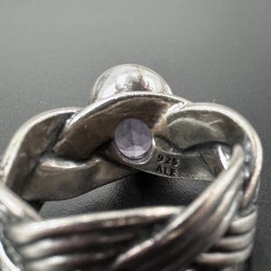 May include: A silver ring with a woven band design. The ring features a small, round, light purple gemstone and a larger, clear, round stone. The ring is stamped with "925 ALE".
