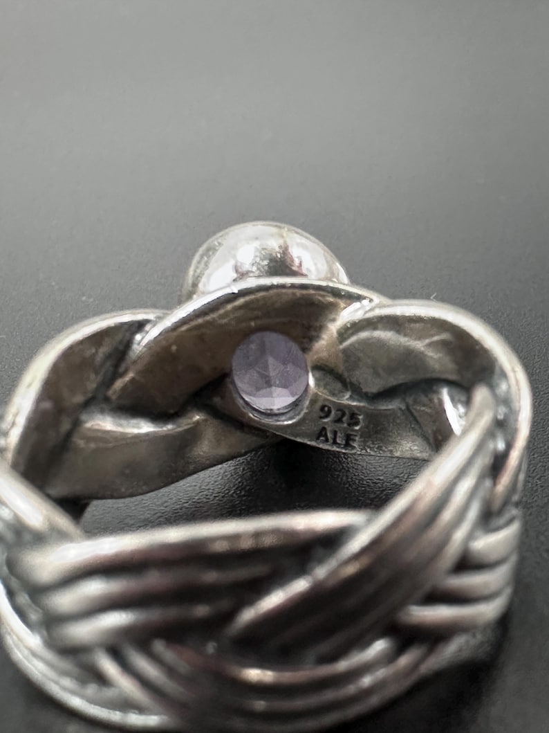 May include: A silver ring with a woven band design. The ring features a small, round, light purple gemstone and a larger, clear, round stone. The ring is stamped with "925 ALE".