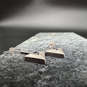 May include: A pair of silver-toned earrings. Each earring features a geometric, triangular design with a textured surface. The earrings are suspended from a post and a small bar. The background is a dark, blurred gradient.