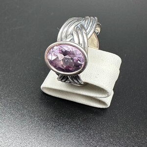 May include: A silver ring featuring a large, oval-cut, light purple gemstone. The band has a woven design, adding texture and visual interest. The ring is displayed on a white surface against a dark background.