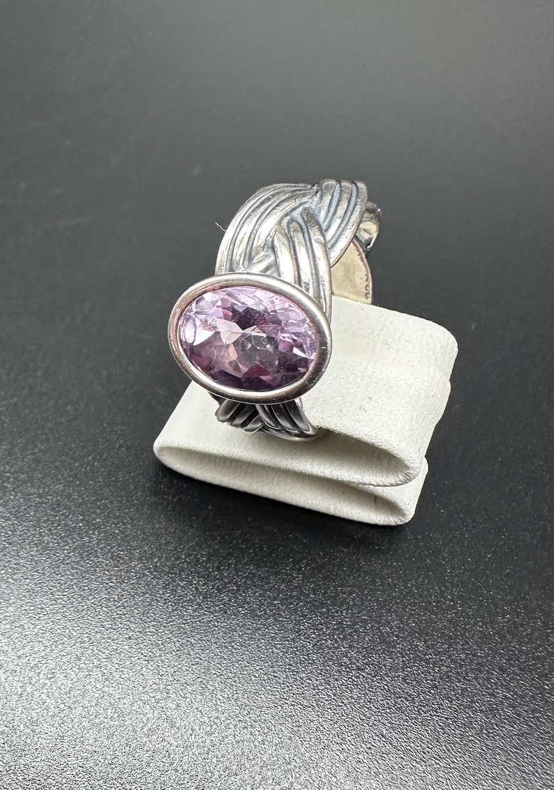 May include: A silver ring featuring a large, oval-cut, light purple gemstone. The band has a woven design, adding texture and visual interest. The ring is displayed on a white surface against a dark background.