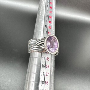 May include: A silver ring with a woven band design and a large oval-shaped purple gemstone. The ring is being measured on a ring sizer, showing the size. The ring is a piece of jewelry.