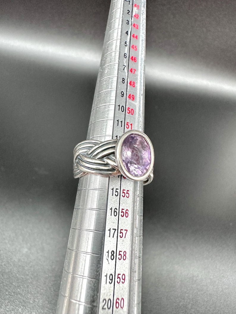 May include: A silver ring with a woven band design and a large oval-shaped purple gemstone. The ring is being measured on a ring sizer, showing the size. The ring is a piece of jewelry.