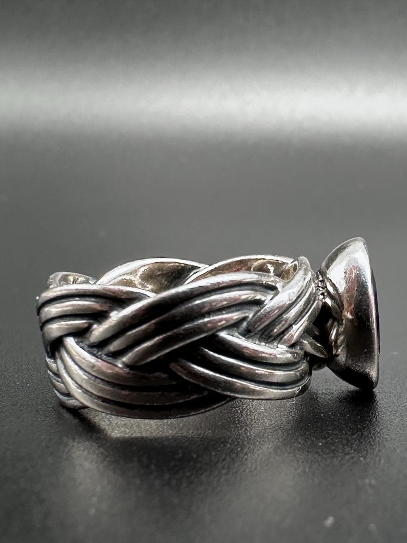 May include: A silver ring with a braided band design and a large, oval, dark purple gemstone. The ring is set against a dark background, highlighting the intricate details of the metalwork and the gemstone's color.