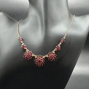 May include: A gold-tone necklace featuring a series of clusters set with deep red garnet-colored stones. The design includes three prominent central floral-shaped clusters flanked by smaller decorative accents along a delicate link chain.