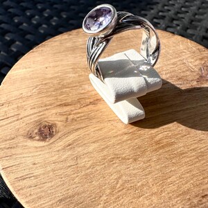 May include: A silver ring with a large, oval, light purple gemstone. The ring's band has a braided design. The ring is displayed on a white stand, set against a wooden surface.