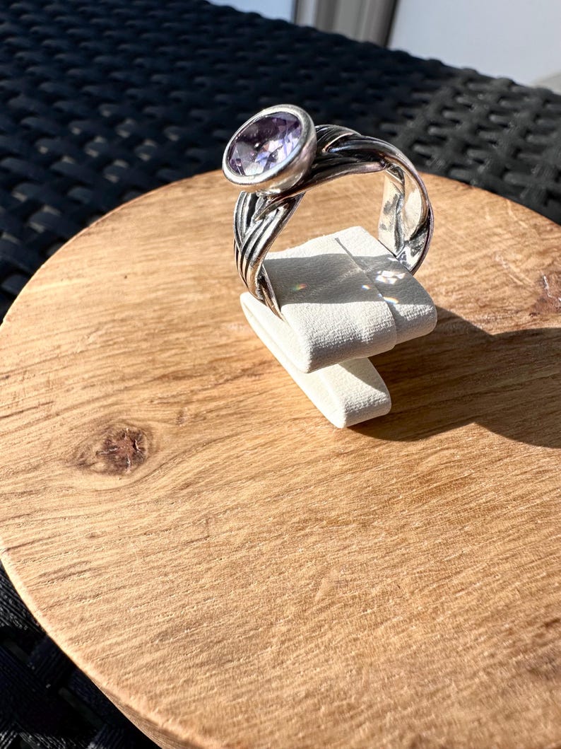 May include: A silver ring with a large, oval, light purple gemstone. The ring's band has a braided design. The ring is displayed on a white stand, set against a wooden surface.