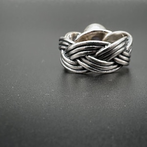May include: A silver ring with a woven band design. The ring features a central, rounded stone. The band has a braided appearance, with multiple strands of metal intertwined to create the woven effect. The ring is set against a dark background.