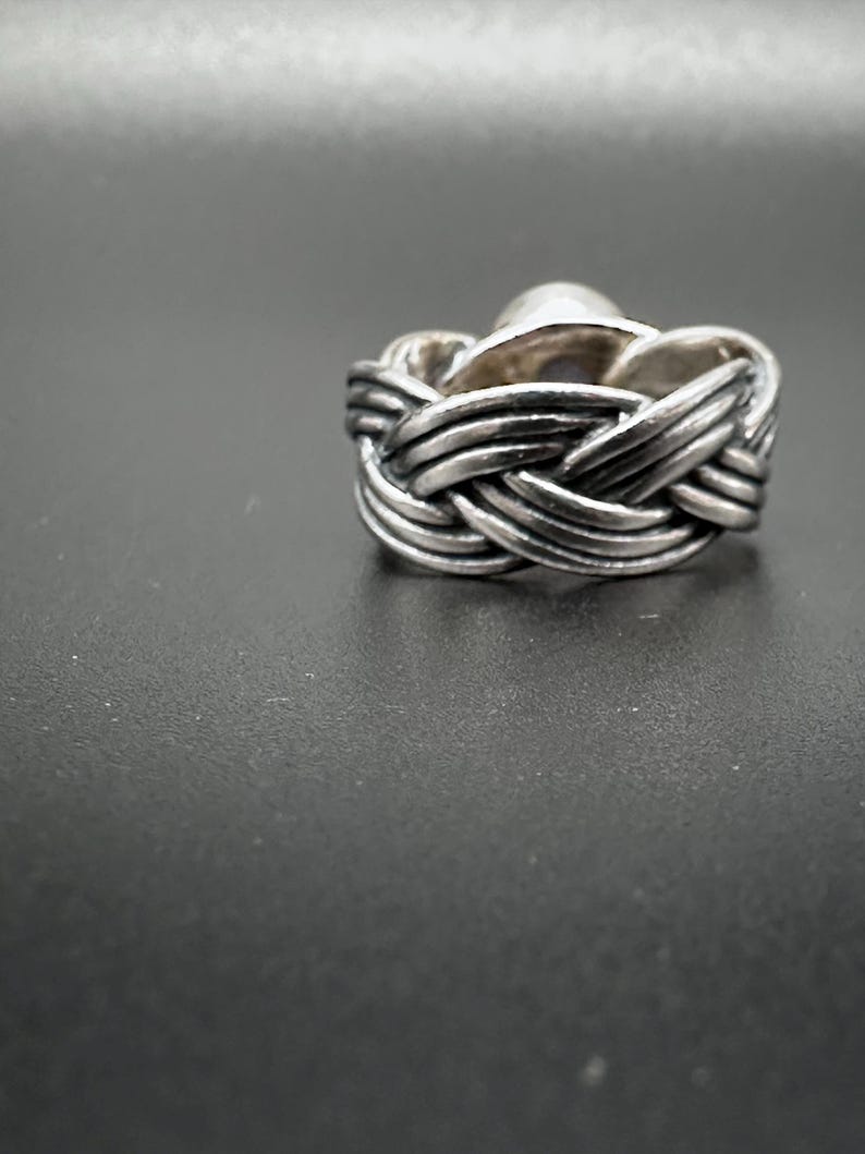 May include: A silver ring with a woven band design. The ring features a central, rounded stone. The band has a braided appearance, with multiple strands of metal intertwined to create the woven effect. The ring is set against a dark background.