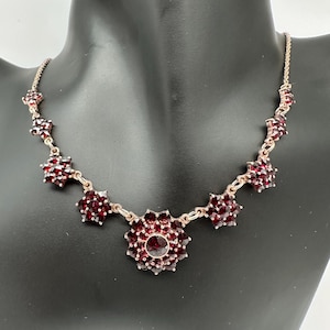 May include: A vintage necklace featuring a series of dark red, flower-shaped garnet clusters. The necklace has a delicate gold-toned chain and a central, larger floral pendant. The garnets are faceted, catching the light and adding sparkle.