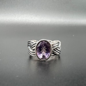 May include: A silver ring with a woven band design. The ring features a large, oval-cut amethyst gemstone in a bezel setting. The amethyst has a light purple hue. The ring is displayed against a dark background.