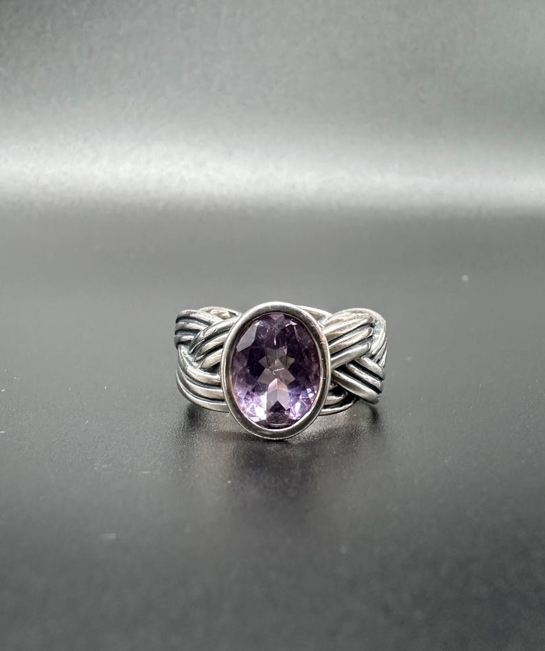 May include: A silver ring with a woven band design. The ring features a large, oval-cut amethyst gemstone in a bezel setting. The amethyst has a light purple hue. The ring is displayed against a dark background.
